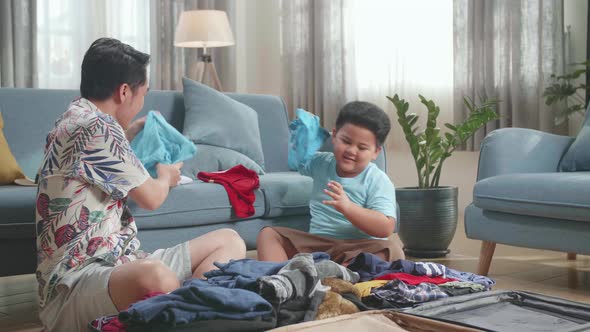 Excited Asian Young Dad And Son Packing Clothes In Suitcase At Home, Preparing For Their Vacation