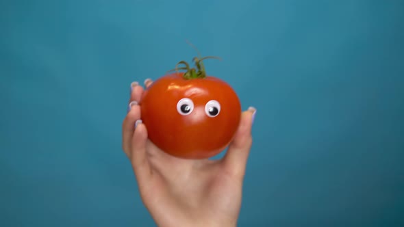 Red Tomato with Eyes in a Woman Hand. Tomato Jumps Into the Frame and Looks Around on a Blue