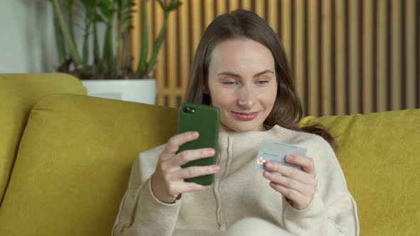 Woman Buying Online with a Credit Card and Smartphone Sitting on a Yellow Couch at Home