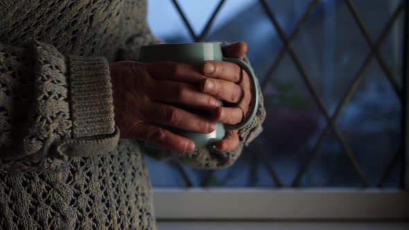 Woman warming hands on hot drink in window medium shot