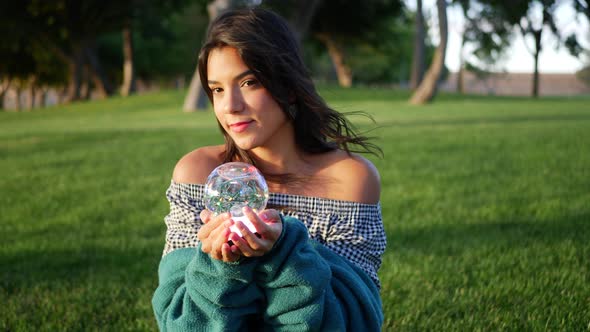 A cute young hispanic woman smiling happily in a park with a magic crystal ball.