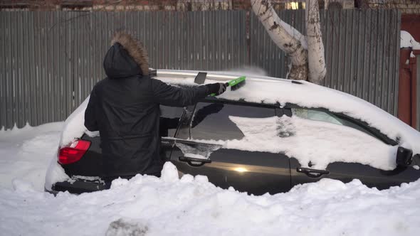 A Young Man in a Jacket Cleans His Car of Snow with a Brush