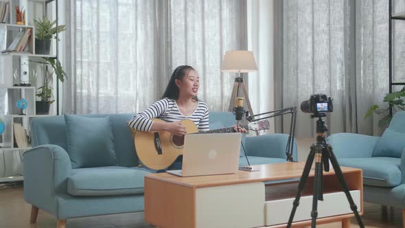 Asian Woman With A Laptop Singing And Playing A Guitar While Live Stream At Home