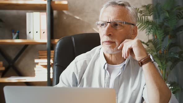 Mature Businessman is Looking Thoughtful Sitting at Desk and Typing on His Laptop While Working at