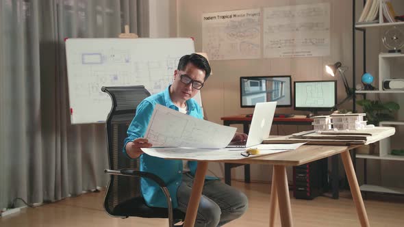 Asian Man Engineer Looking At Blueprint And Typing On A Laptop At The Office alt
