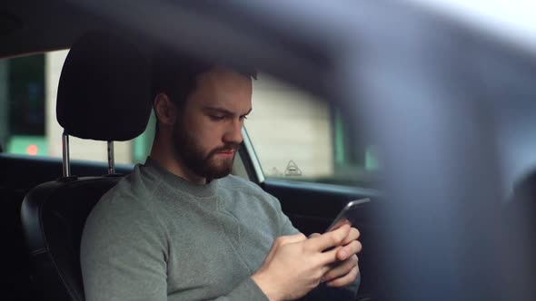 Tracking shot of serious handsome young man sitting in car, looking on display screen of cell phone