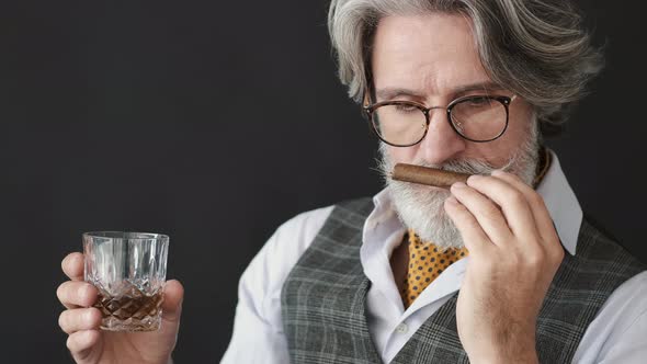 Mature Man with a Glass of Whiskey Smelling Cigar Over Black Background