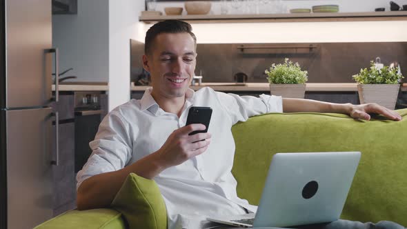 Young Happy Male Entrepreneur Uses Smartphone While Working at a Laptop Sitting at Home on the Couch