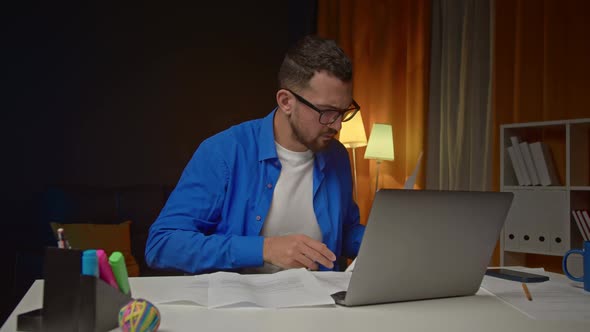 Bearded Man in White Tshirt Working with Laptop and Papers at Home