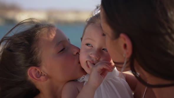 Closeup Portrait of Carefree Pretty Baby Girl with Cheerful Sisters Kissing Toddler on Cheeks in