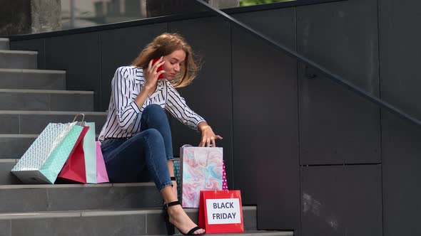 Girl Sitting on Stairs with Bags Talking on Smartphone About Sale in Shopping Mall in Black Friday