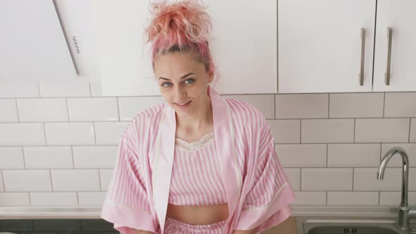 Young Beautiful Girl with Pink Hair in Pink Pajamas Drinking Tea in White Loft Kitchen