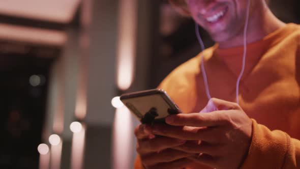 Caucasian male wearing earphones, smiling, using his phone in a street the evening