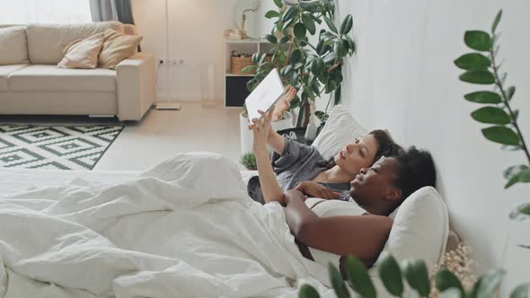 Cheerful Queer Couple with Tablet Relaxing in Bed