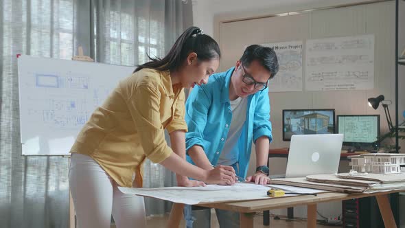 Asian Woman And Man Engineers With Blueprint Discuss About Design At The Office