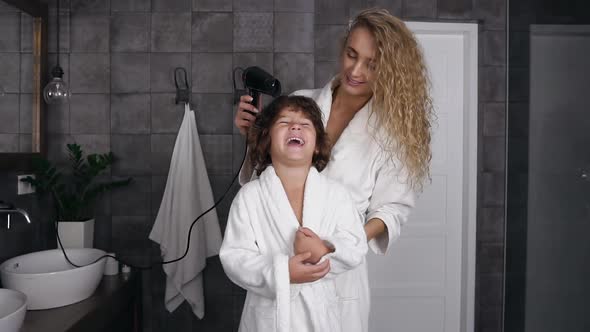 Young Woman with Long Curly Hair Dries Curly Hair with a Hairdryer Her Little Happy Son