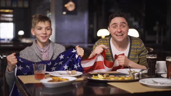 Excited Caucasian Man and Teenage Boy Rejoicing Shaking American Flag Dining in Restaurant Watching