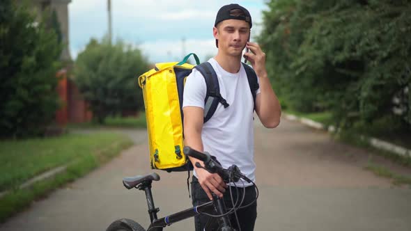 Caucasian Courier Talking on Phone with Client Standing on Suburban Road with Bike and Yellow