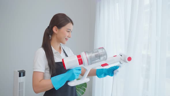 Asian cleaning service woman worker clean in living room and vacuuming messy dirty curtain at home.