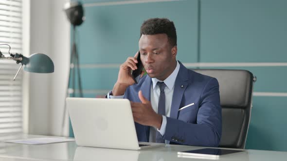 Angry Businessman Talking on Smartphone While Using Laptop in Office