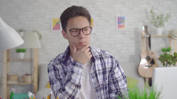 Thoughtful Young Asian Man in Stylish Glasses Working at Laptop in Living Room at Home