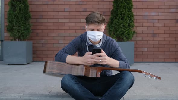 Young Guitarist Is Sitting on the Sidewalk and Typing a Message To His Friends