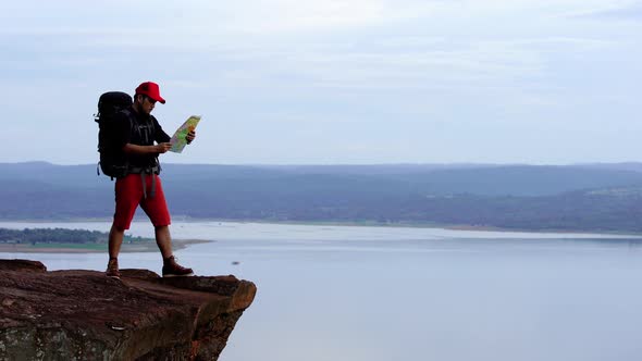 man traveler with backpack looking at the map on the edge of cliff, on a top of the rock mountain