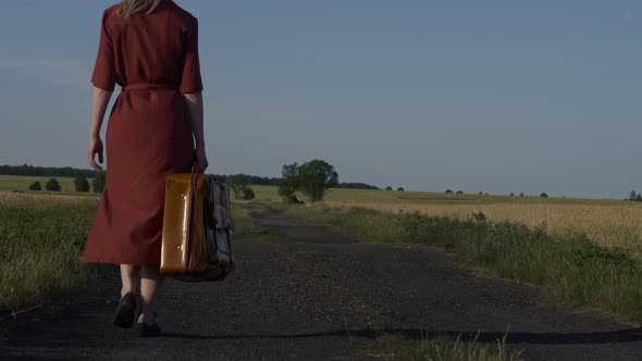 Girl in red dress with suitcase on country road in sunset.