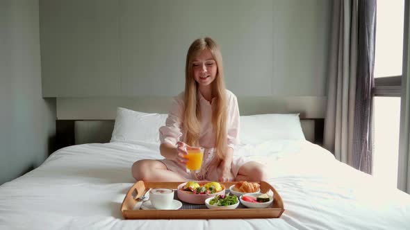 Young Smiling Beautiful Woman Eating Breakfast in Bed in Cozy Hotel Room