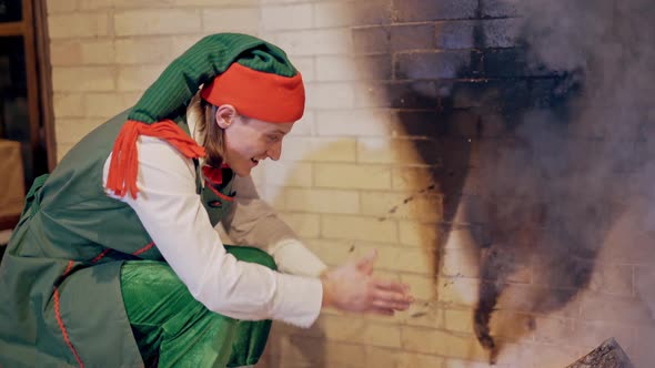 Elf is sitting near the fire and warming his cold hands in the house. 