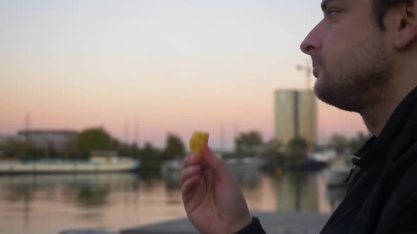 Close up of a young man taking an orange slice and eating it on a pier during sunset