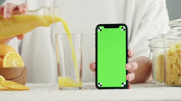 Woman Pouring Orange Juice and Holding Smartphone with Chroma Key Fresh Juice on Kitchen Table