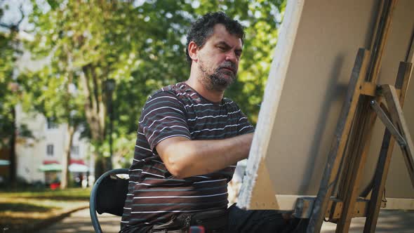 Guy Artist is Drawing a Picture on Canvas Using Oil Paints While Sitting on an Alley of City Park