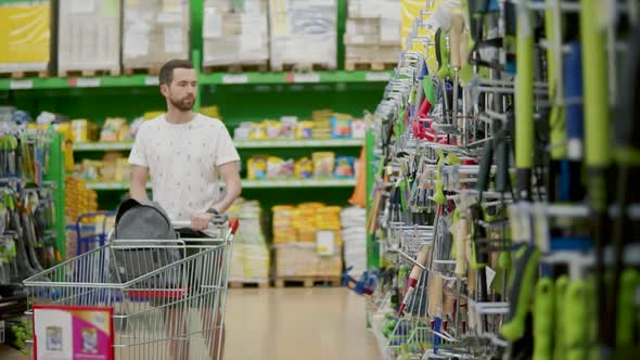 Alone Young Man is Walking in Sales Hall in Shop Rolling Trolley in Front