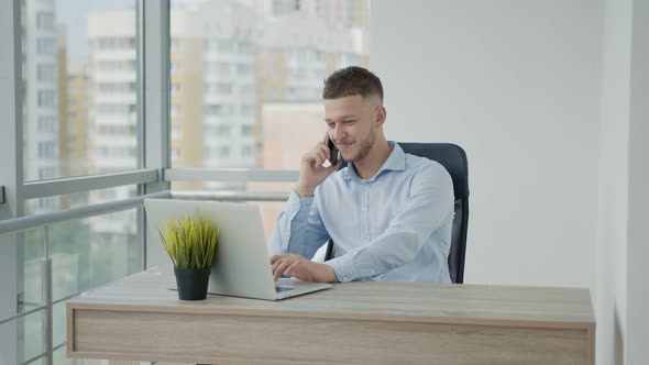 Young Man of Caucasian Appearance Emotionally Talking on the Phone in the Office Sitting at a Table