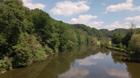 The Sieg flowing calmly in between lush, leafy riverbanks underneath a blue sky with cumulus clouds.