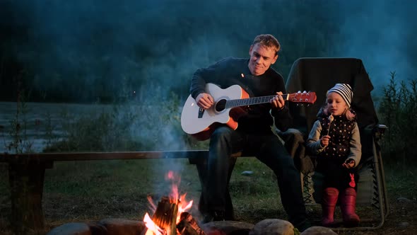 Father Playing Guitar for Daughter Over Campfire