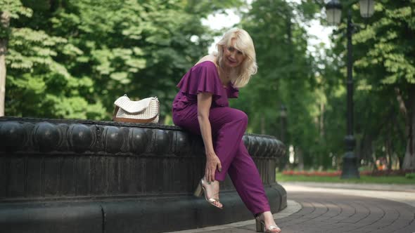 Wide Shot Adult Tired Woman Rubbing Ankle Sitting at Fountain in Summer Park