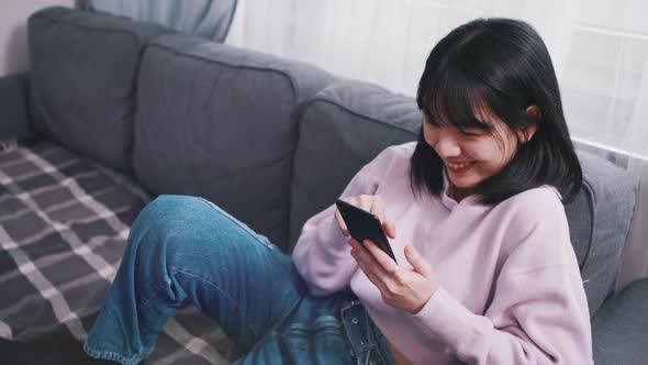 Young Asian Woman Relaxing on the Couch and Smiling While Reading Message on the Smartphone