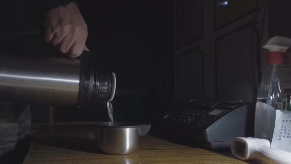 Man Pours Hot Tea From a Thermos Into a Mug on a Table in a Dark Room on Night Duty