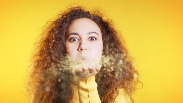 Attractive Girl Blowing Shiny Dust or Sparkles Confetti on Yellow in Studio