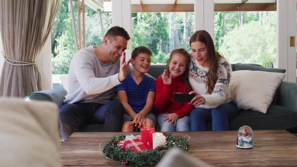 Caucasian family smiling and waving while having a video call on smartphone sitting on the couch at