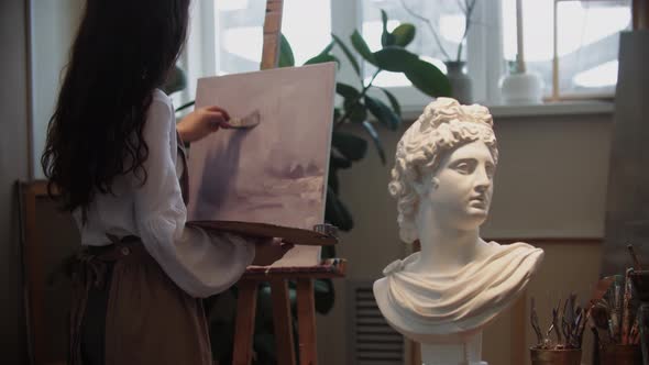 Young Woman Artist with Beautiful Hair Working with a Painting  Marble Bust on the Foreground