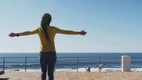 African american woman walking with arms outstretched on promenade by the sea