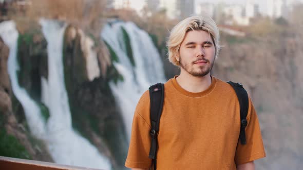 A Young Blonde Man Stands on the Background of Waterfall