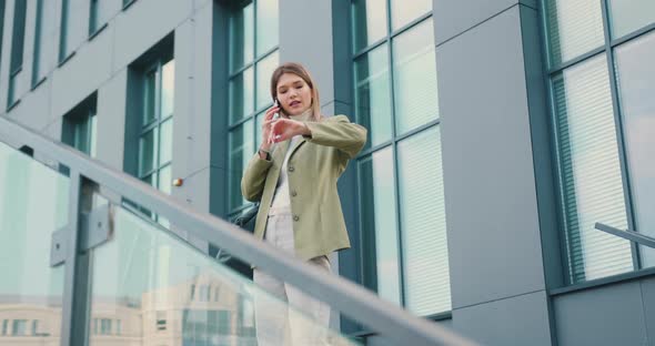 Smiling Woman Looking on Watch While Talking on Smartphone During Walking in Urban City