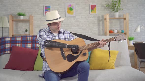 Young Man in a Hat with a Bionic Prosthetic Arm Plays an Acoustic Guitar Sitting on a Sofa