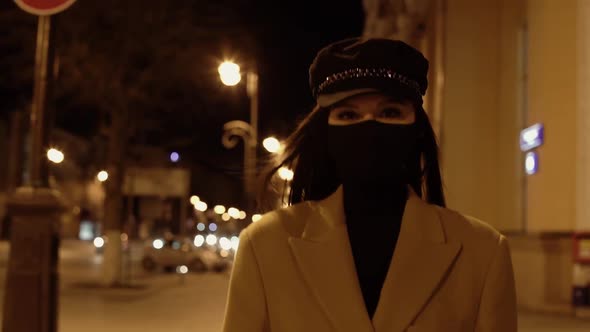 Girl in a Black Virus Mask in a Beige Coat Crosses the Road in the Night City