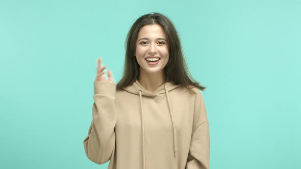 Attractive Caucasian Girl in Beige Hoodie Waving Hand and Saying Hello Blink Eyes and Glancing