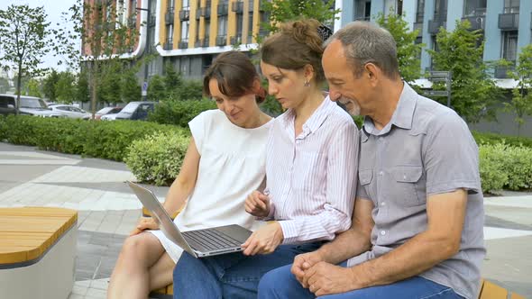 Senior Businessman and Two Female Coworkers are Discussing the Project Sitting on the Bench Near New